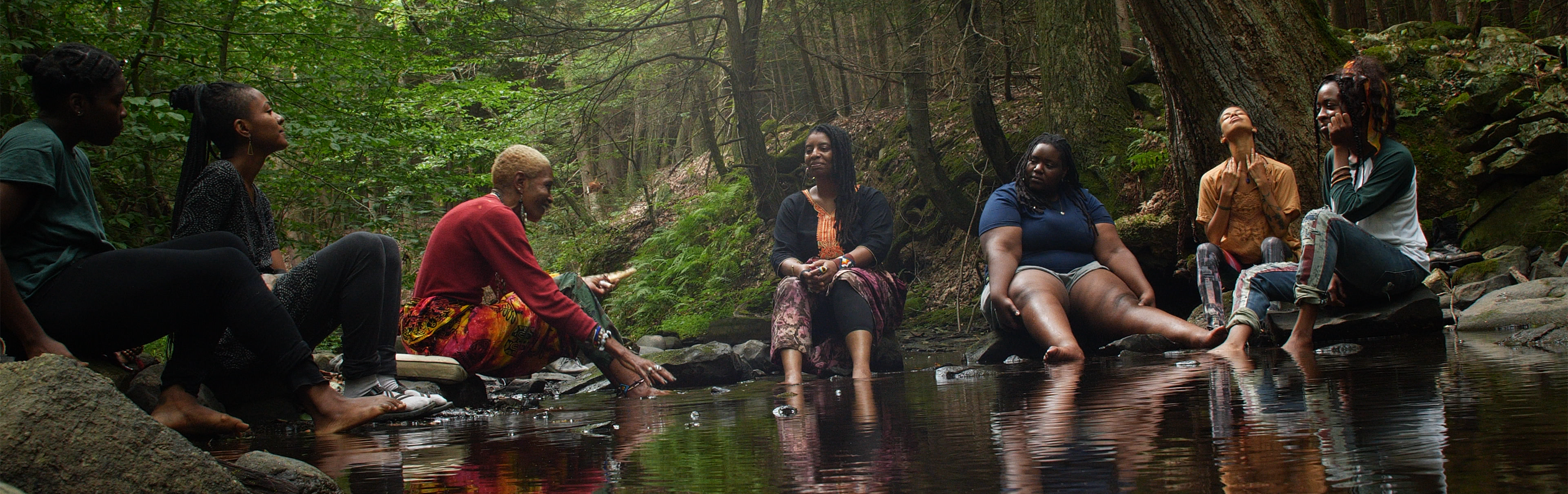 Six black women sit around on the banks of a creek with their feet in the water, singing.