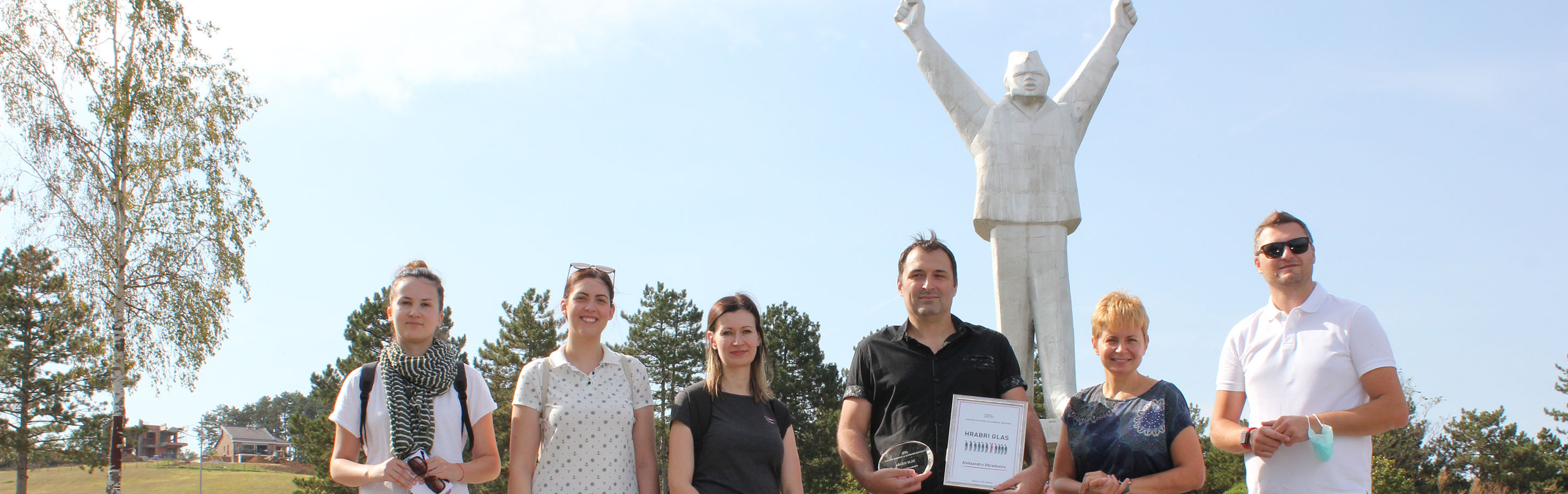 A man holding a trophy, flanked by colleagues, poses in front of a marble statue of a man holding his arms up in victory.