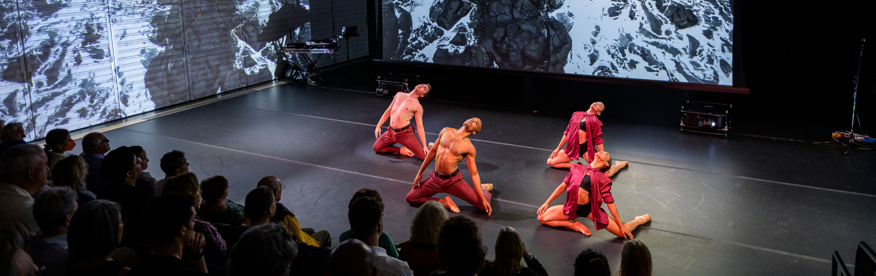 Dancers in deep red costumes appear on their knees on stage in front of an audience against a background of the ocean. 