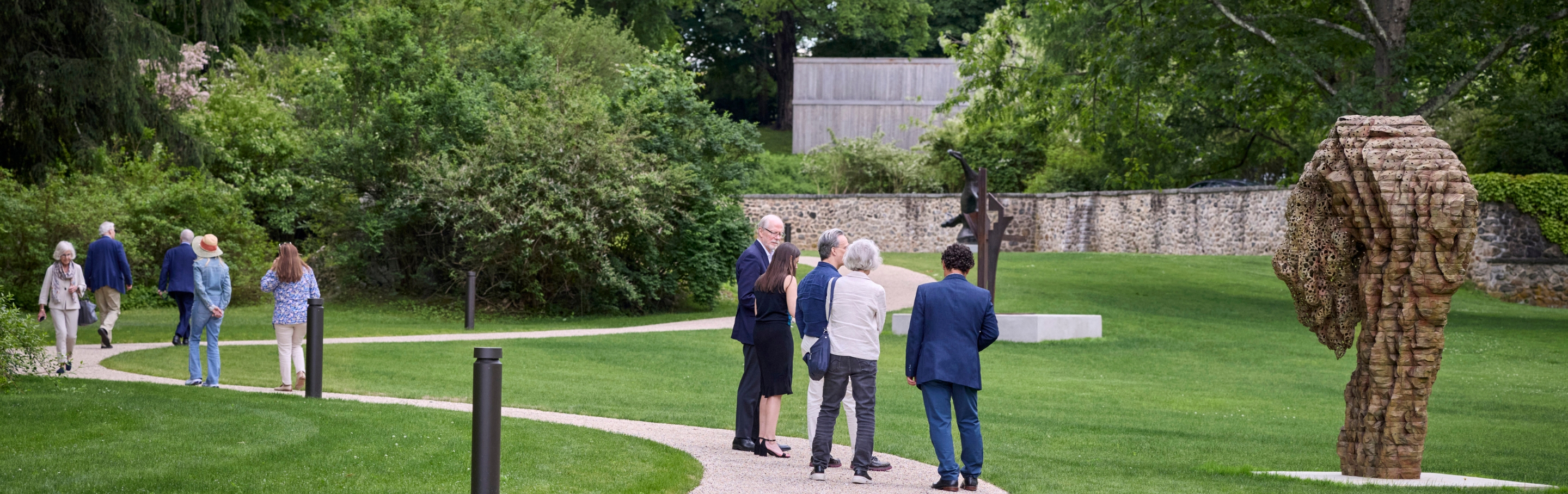 Visitors stroll along a winding gravel path through a landscaped sculpture garden, pausing to view large outdoor sculptures set on a manicured lawn surrounded by mature trees and shrubs.