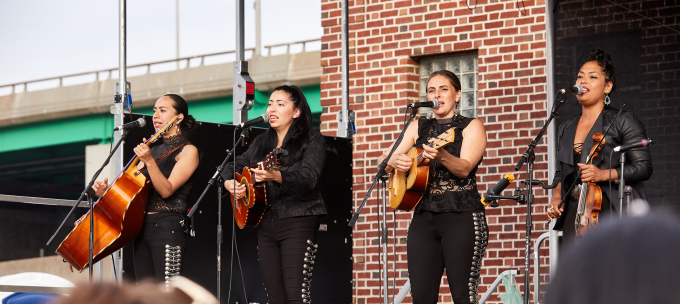An all-female mariachi bands performs on stage for a live audience.