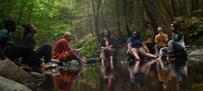Six black women sit around on the banks of a creek with their feet in the water, singing.