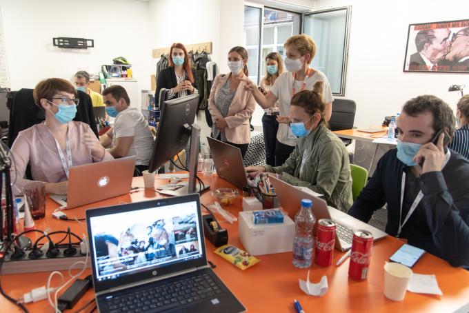 a crowd of masked workers around a table, some conversing, others on their phones or computers.