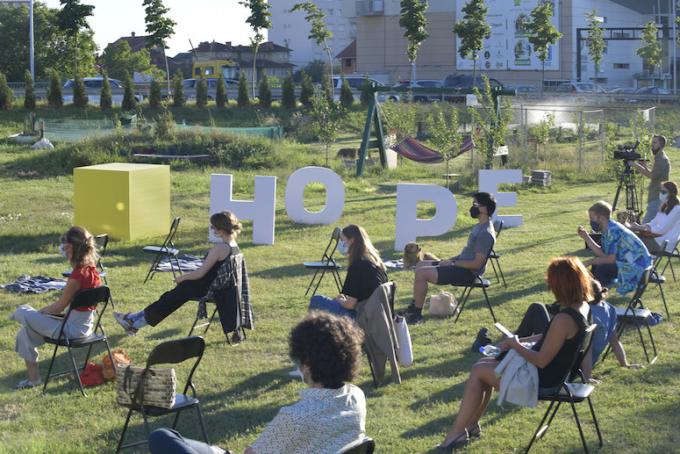 audiences seated outdoors, masked, and separated by 6 feet outside, with a stature of the word hope in the background.