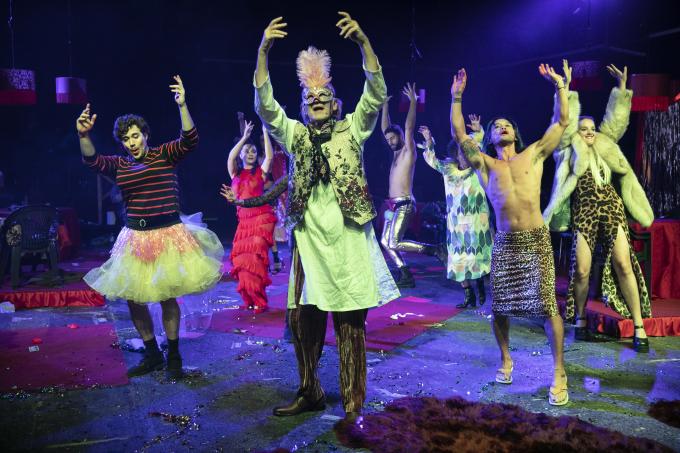 A cast of brightly costumed, masked performers dance under moody stage lights.