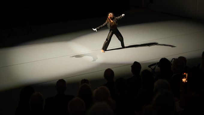 a solo dancer performs on a starkly lit stage before an audience