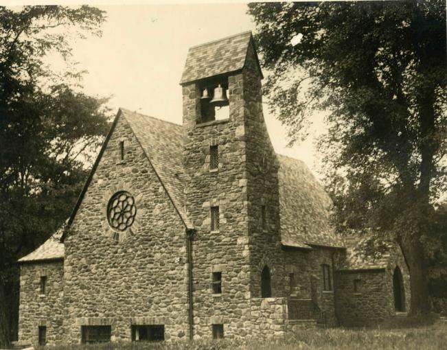 Black and white photo of the exterior of a stone church featuring a circular stained glass window and a stone belfry.