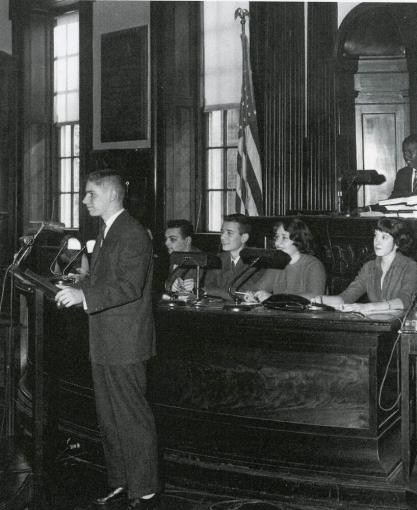 A young man stands at a lectern in a courtroom.