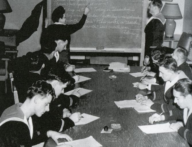 Men wearing naval uniforms sit at a table in a classroom with a chalkboard.