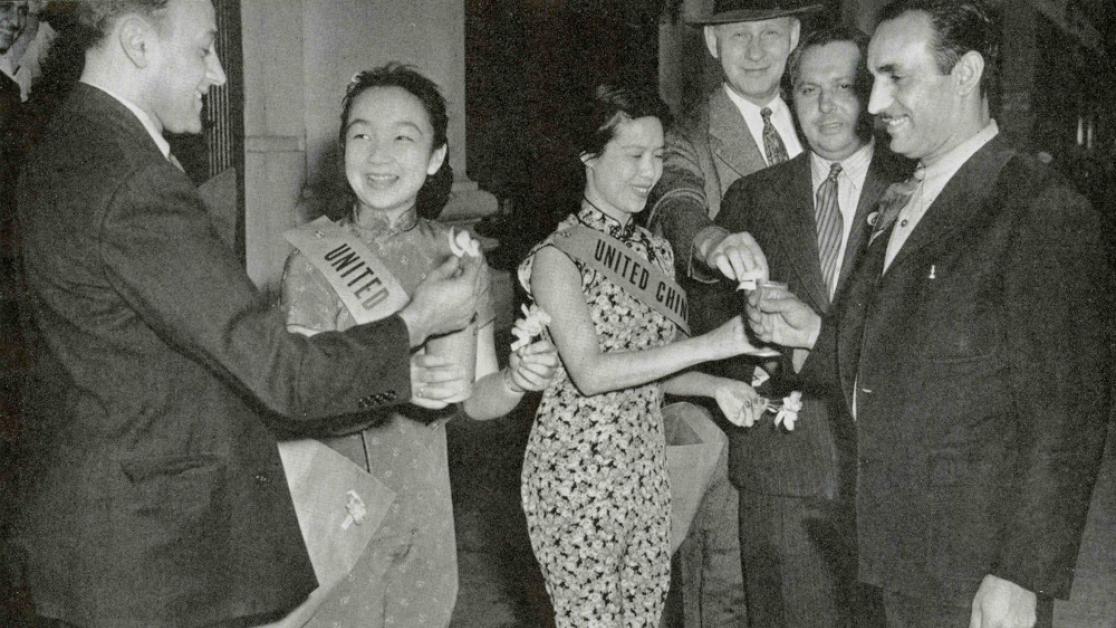 Two women wearing dresses stand in between several men wearing suits.