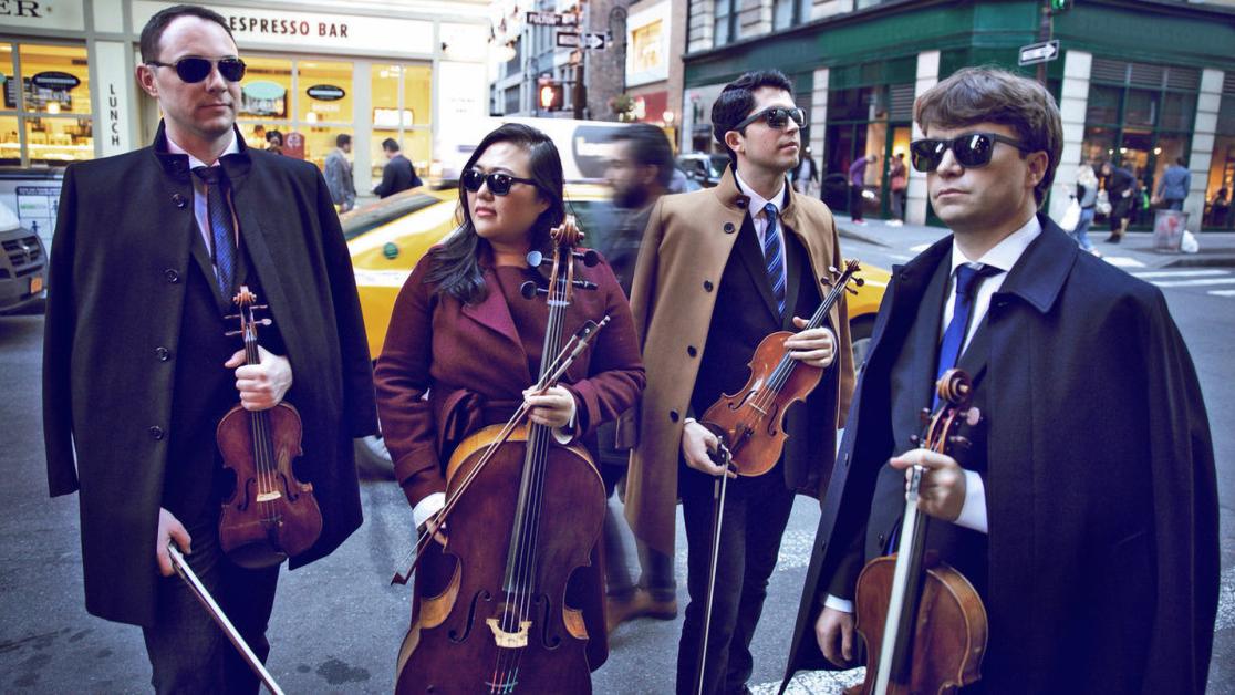 Four string quartet musicians wearing sunglasses hold their instruments on a busy New York City street.