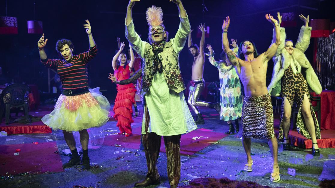 A cast of brightly costumed, masked performers dance under moody stage lights.