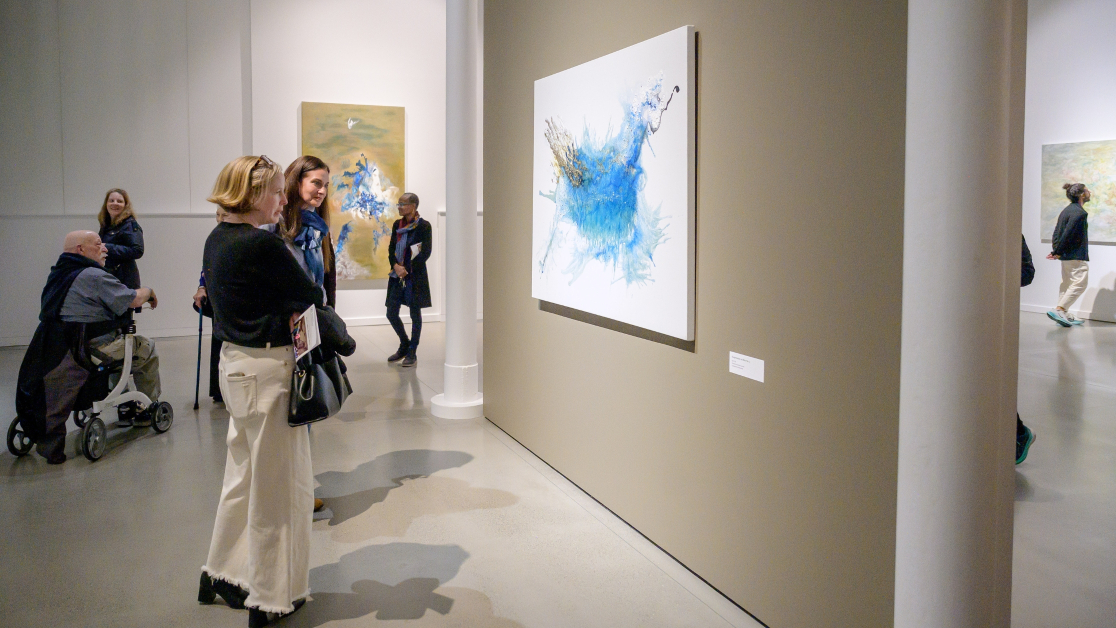 Visitors stand in a contemporary art gallery, looking at large abstract paintings on neutral-colored walls. In the foreground, two women face a blue-and-white abstract artwork, while other visitors—including a person using a mobility scooter—move through the open, well-lit exhibition space.