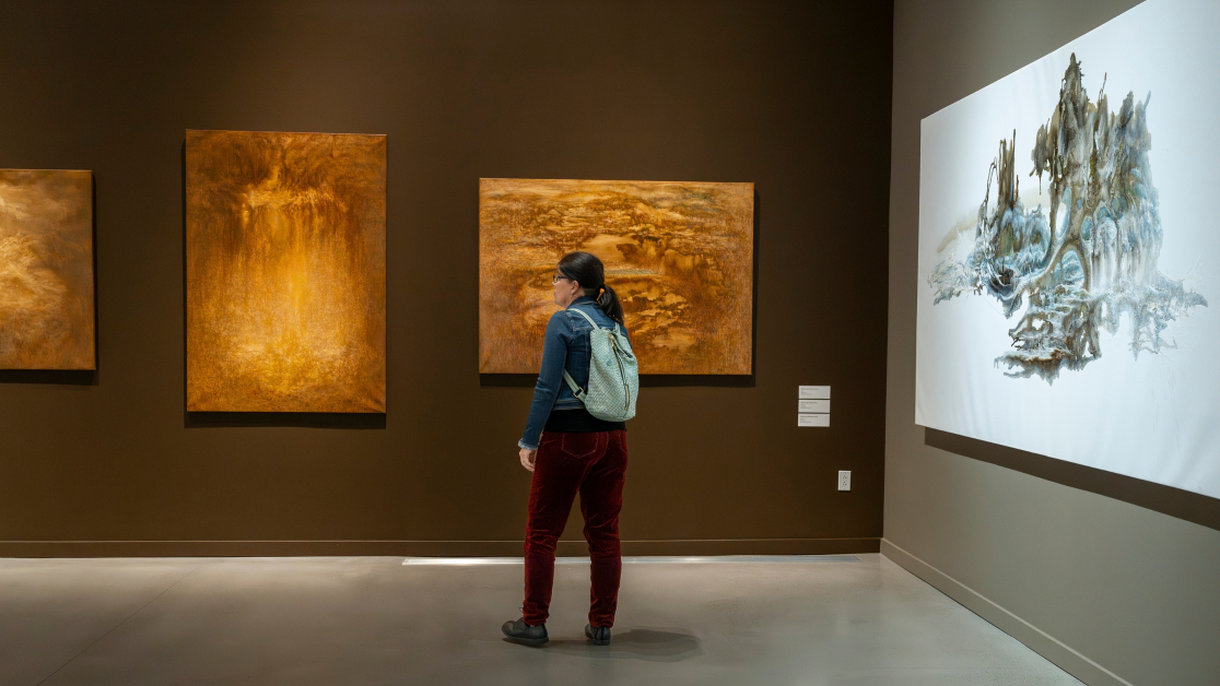 A person wearing a backpack looks at several landscape paintings in an art gallery.