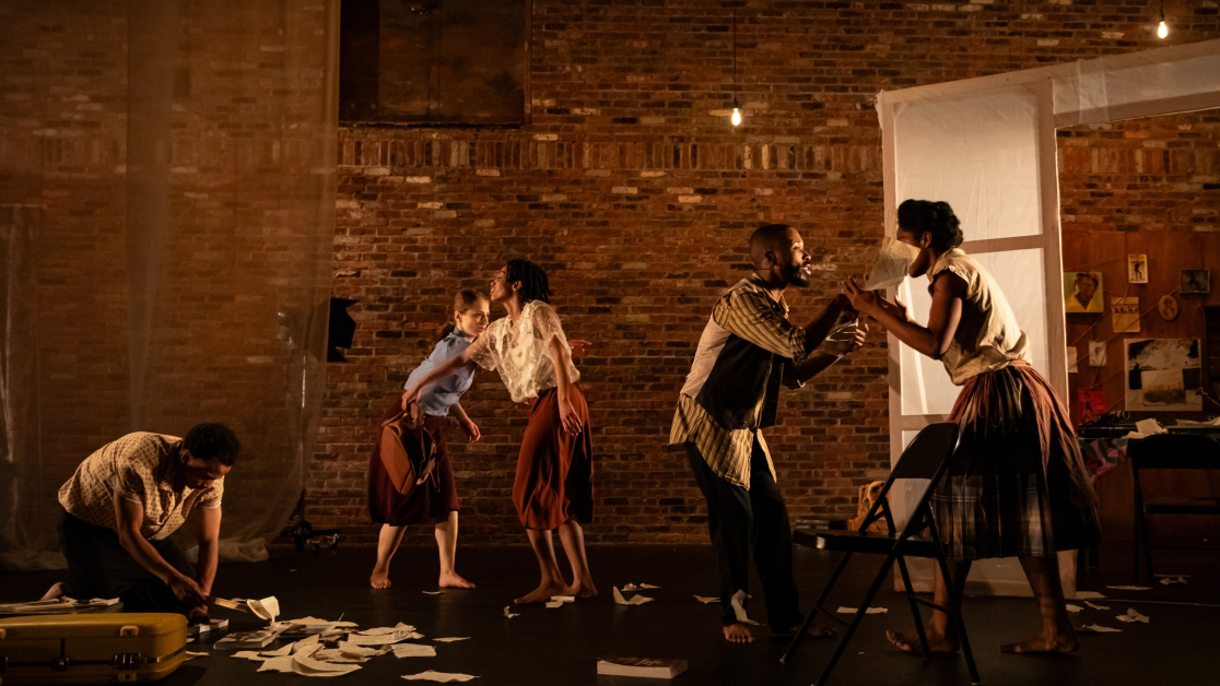 “Five dancers perform on a dimly lit stage with brick walls, scattered papers on the floor, and gestures suggesting tension and interaction.”