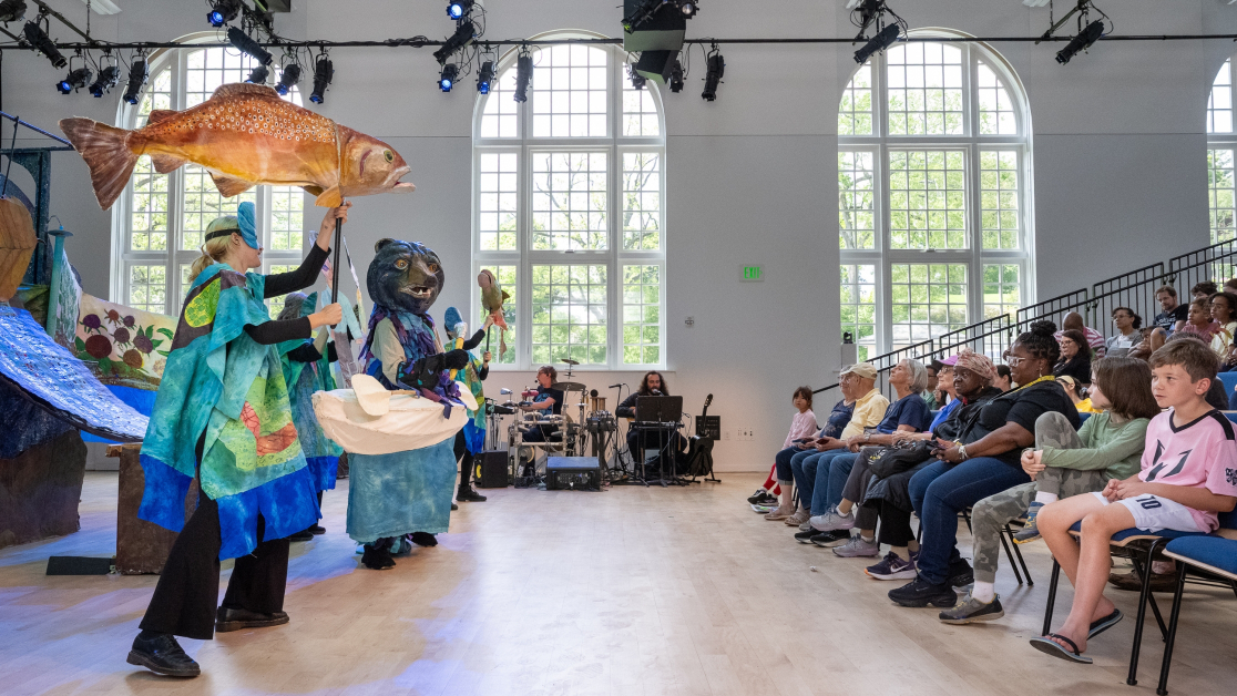 Costumed performers with fish and ocean-themed puppets entertain a seated audience.