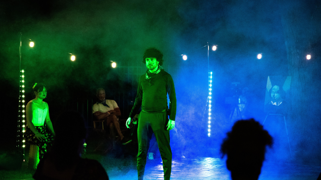 Kamran Saliani stands in a haze of blue and green stage lighting during an outdoor theatrical performance, while audience members and fellow performers look on.