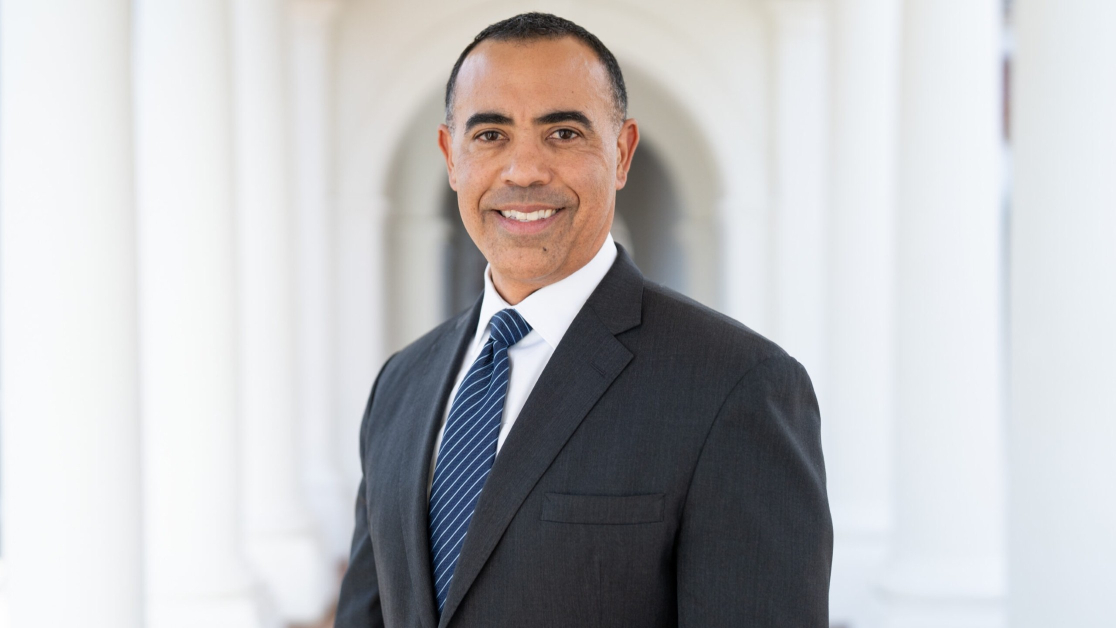 Portrait of Ian Solomon smiling at the camera in a dark gray suit, white shirt, and blue striped tie, standing in a sunlit colonnade with softly blurred arches behind him. 