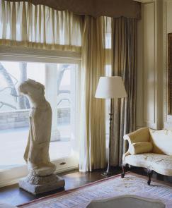 a brightly light room displays a a headless Bodhisattva sculpture, a portrait of Abby Aldrich Rockefeller, and an cream antique couch.