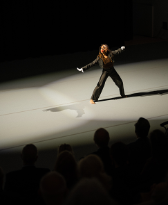 a solo dancer performs on a starkly lit stage before an audience