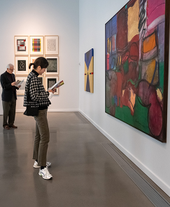 Two visitors examine colorful abstract paintings hanging in a gallery.