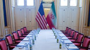 An American flag and an Iranian flag at the end of a long table set with empty chairs on either side of it.