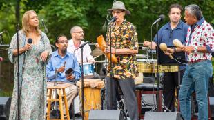 Members of a band on stage playing various instruments including drums and maracas.