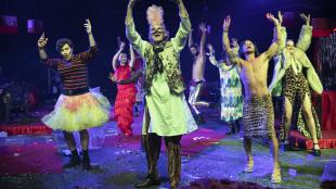 A cast of brightly costumed, masked performers dance under moody stage lights.