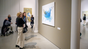 Visitors stand in a contemporary art gallery, looking at large abstract paintings on neutral-colored walls. In the foreground, two women face a blue-and-white abstract artwork, while other visitors—including a person using a mobility scooter—move through the open, well-lit exhibition space.