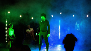 Kamran Saliani stands in a haze of blue and green stage lighting during an outdoor theatrical performance, while audience members and fellow performers look on.