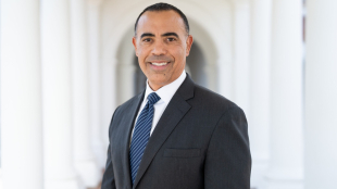Portrait of Ian Solomon smiling at the camera in a dark gray suit, white shirt, and blue striped tie, standing in a sunlit colonnade with softly blurred arches behind him. 
