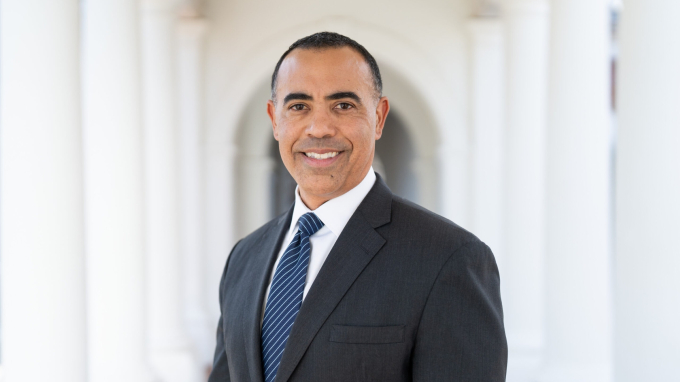 Portrait of Ian Solomon smiling at the camera in a dark gray suit, white shirt, and blue striped tie, standing in a sunlit colonnade with softly blurred arches behind him.