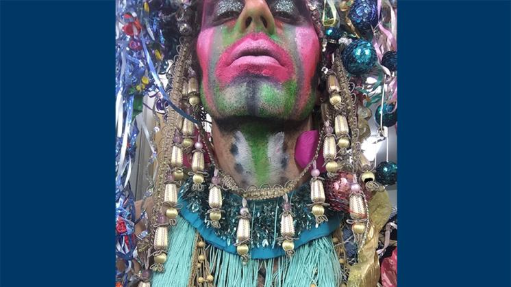 A close-up photo of Machine Dazzle with his face painted with multiple hues and glitter on his eyelids, surrounded by reflective orbs, tassles, and beads.