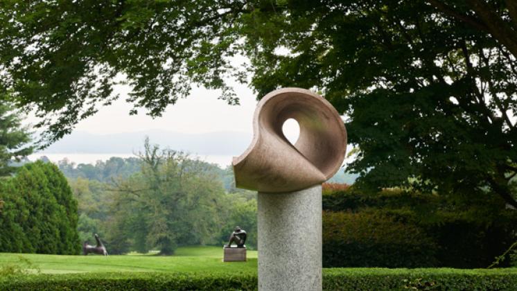 A sculpture sits in the foreground with picturesque green scenery and an additional sculpture in the background.